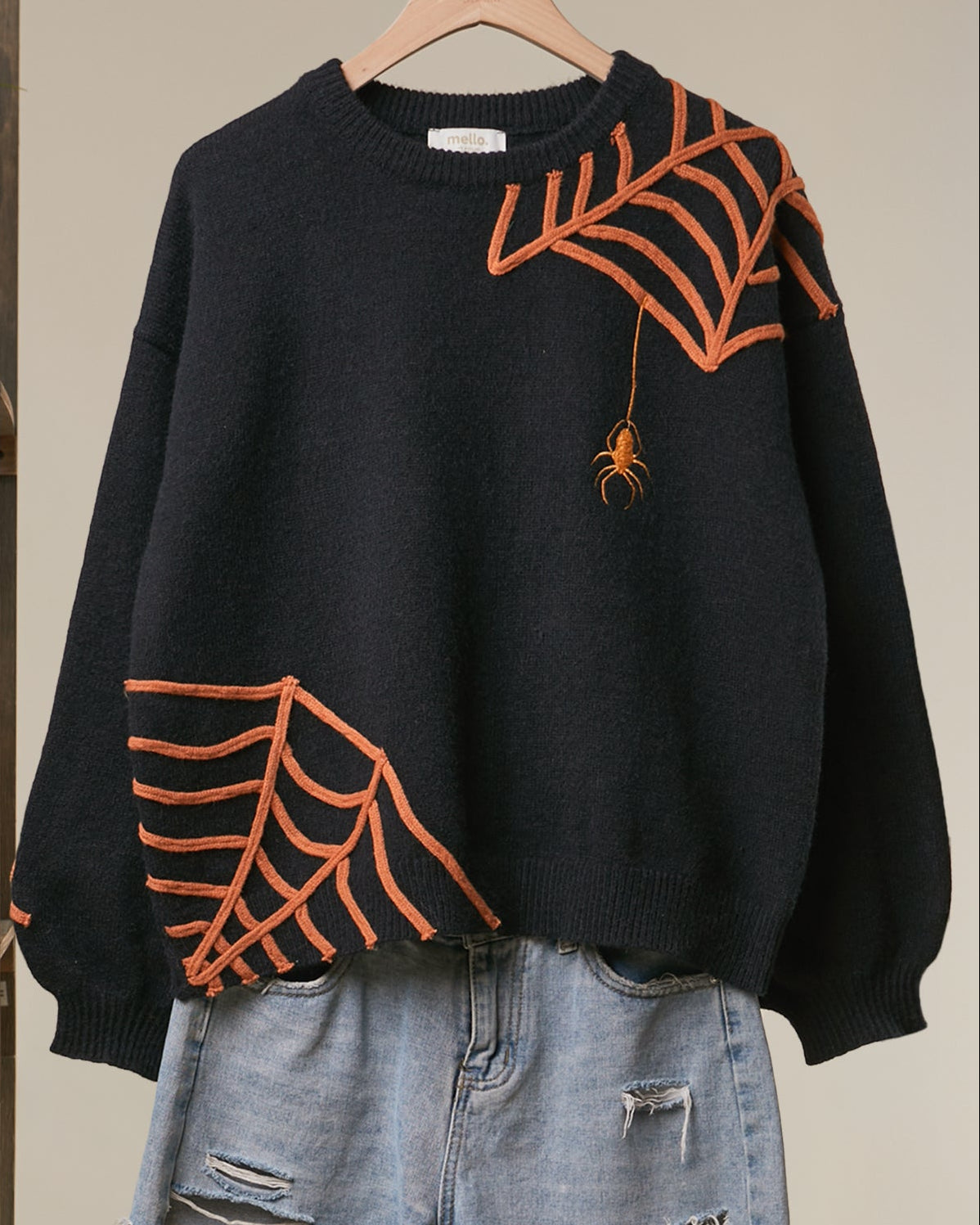 A black crewneck sweater with orange spiderweb and spider motifs, styled with light-wash denim shorts on a wooden hanger against a beige wall.